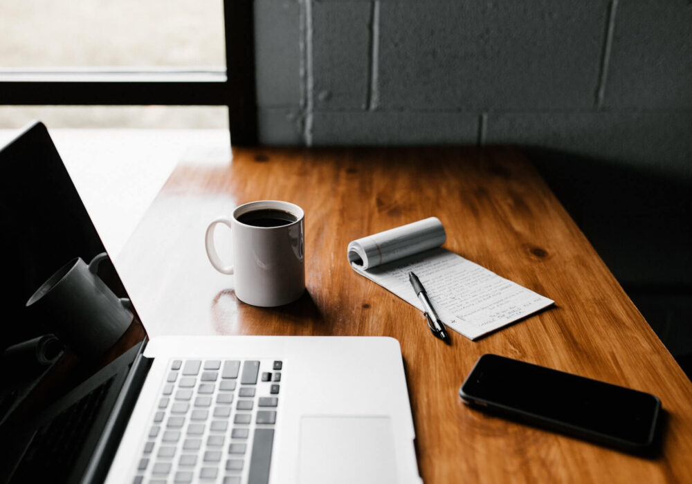 Laptop, a white mug, smartphone and a pen and paper on a table