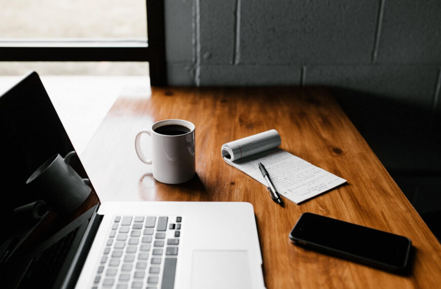 Laptop, a white mug, smartphone and a pen and paper on a table